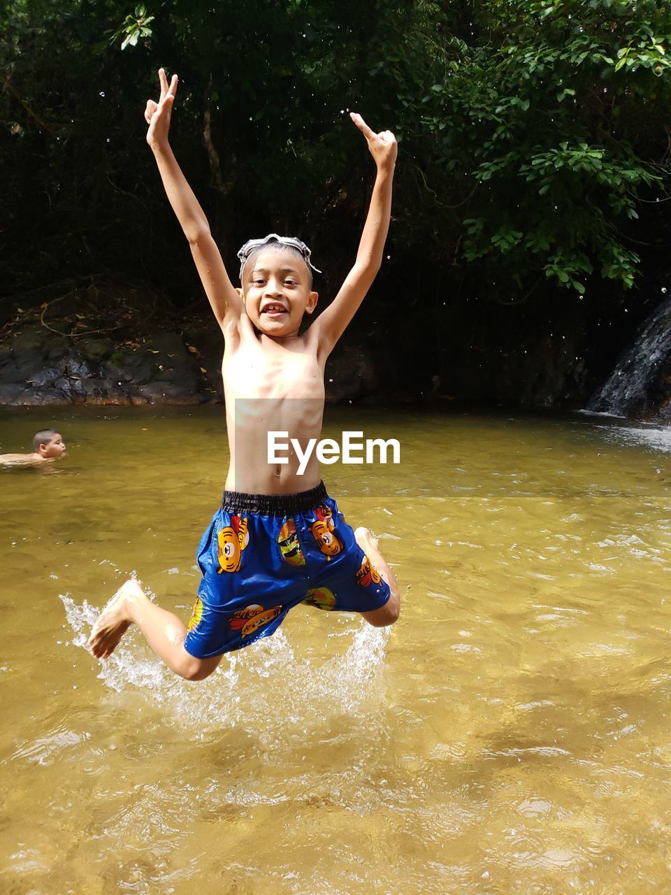 Portrait of shirtless boy with arms raised jumping in river against plants
