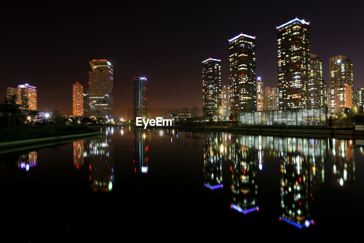 Illuminated buildings by river against sky at night