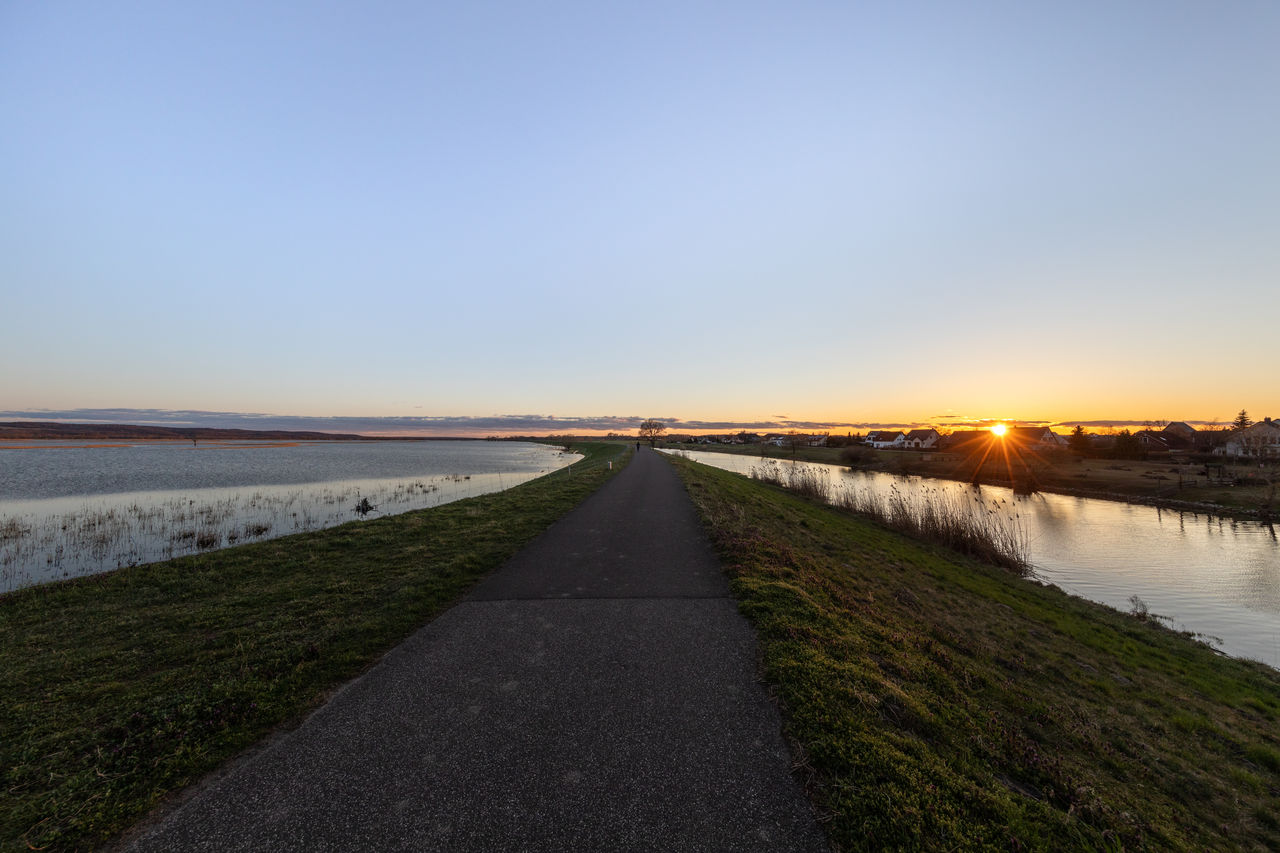 Path on a dam between river and floodplain against clear sky during sunset