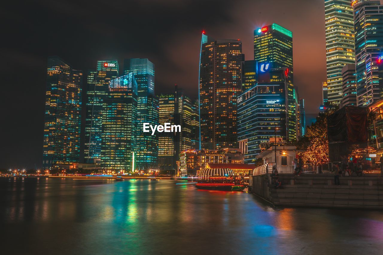 ILLUMINATED BUILDINGS BY RIVER AGAINST SKY