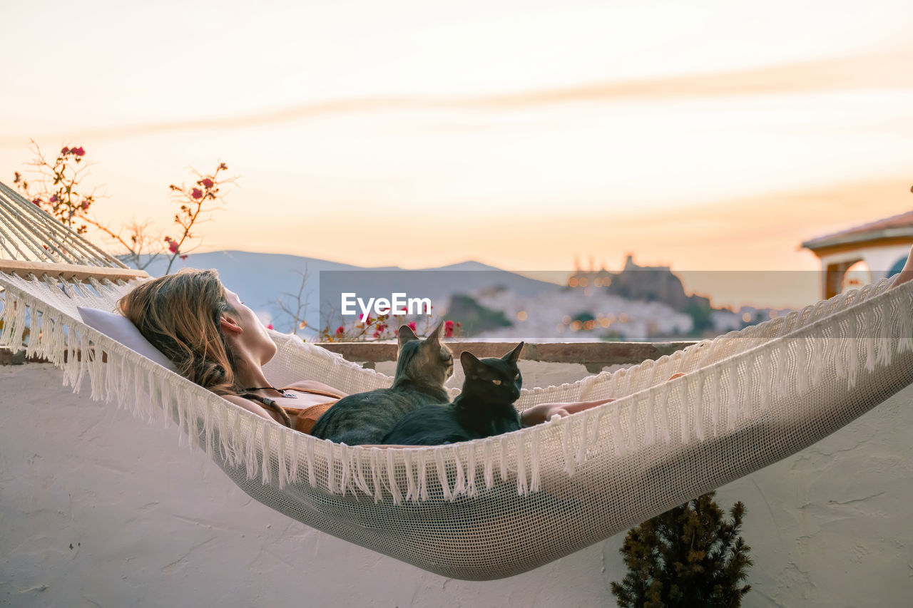 Woman with cats relaxing on hammock against sky during sunset