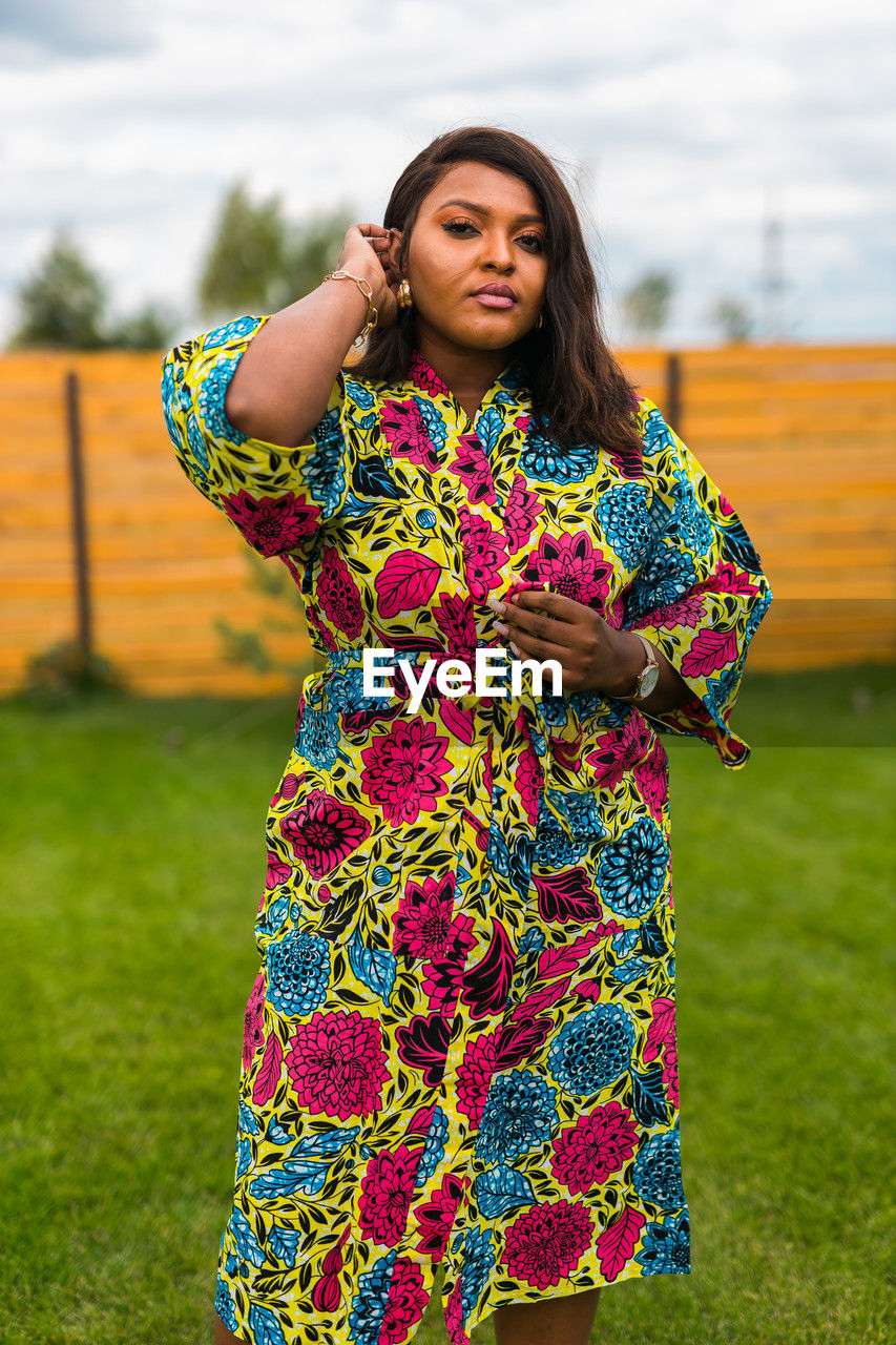 one person, women, dress, adult, clothing, standing, portrait, young adult, smiling, grass, nature, front view, multi colored, happiness, hairstyle, floral pattern, fashion, emotion, yellow, spring, pattern, looking at camera, plant, lifestyles, long hair, outdoors, sky, traditional clothing, brown hair, female, three quarter length, cheerful, person, leisure activity, human face, day, flower, photo shoot, looking, focus on foreground, holding