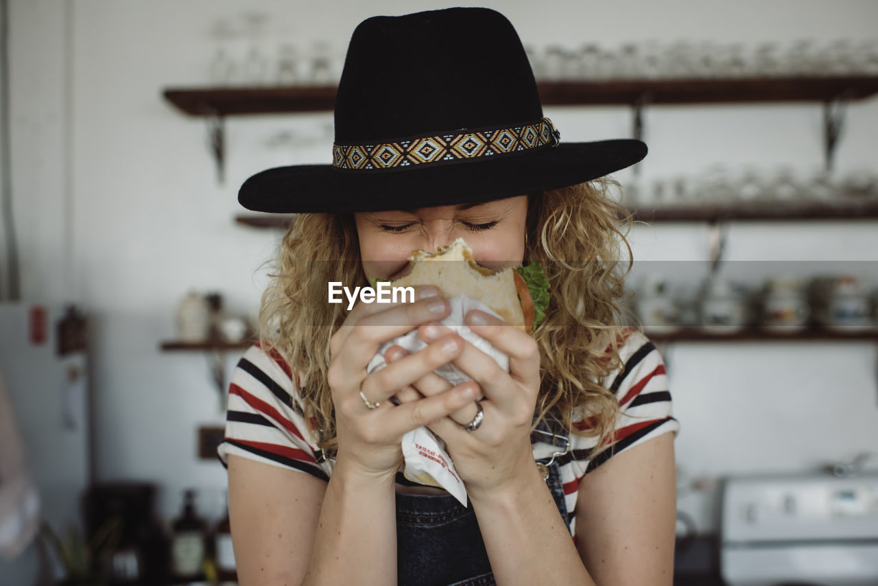 Close-up of woman wearing hat holding sandwich while standing at home