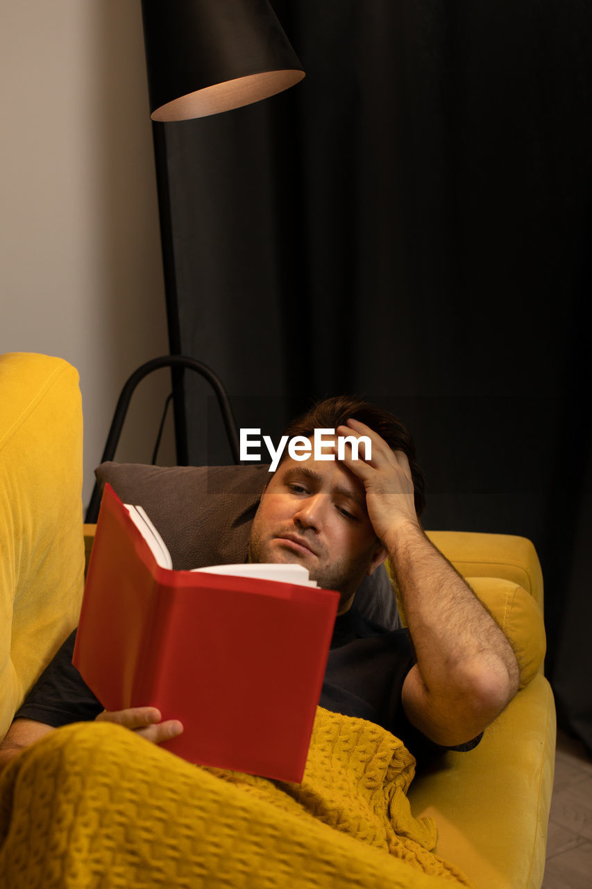 Man reading book while lying on sofa at home