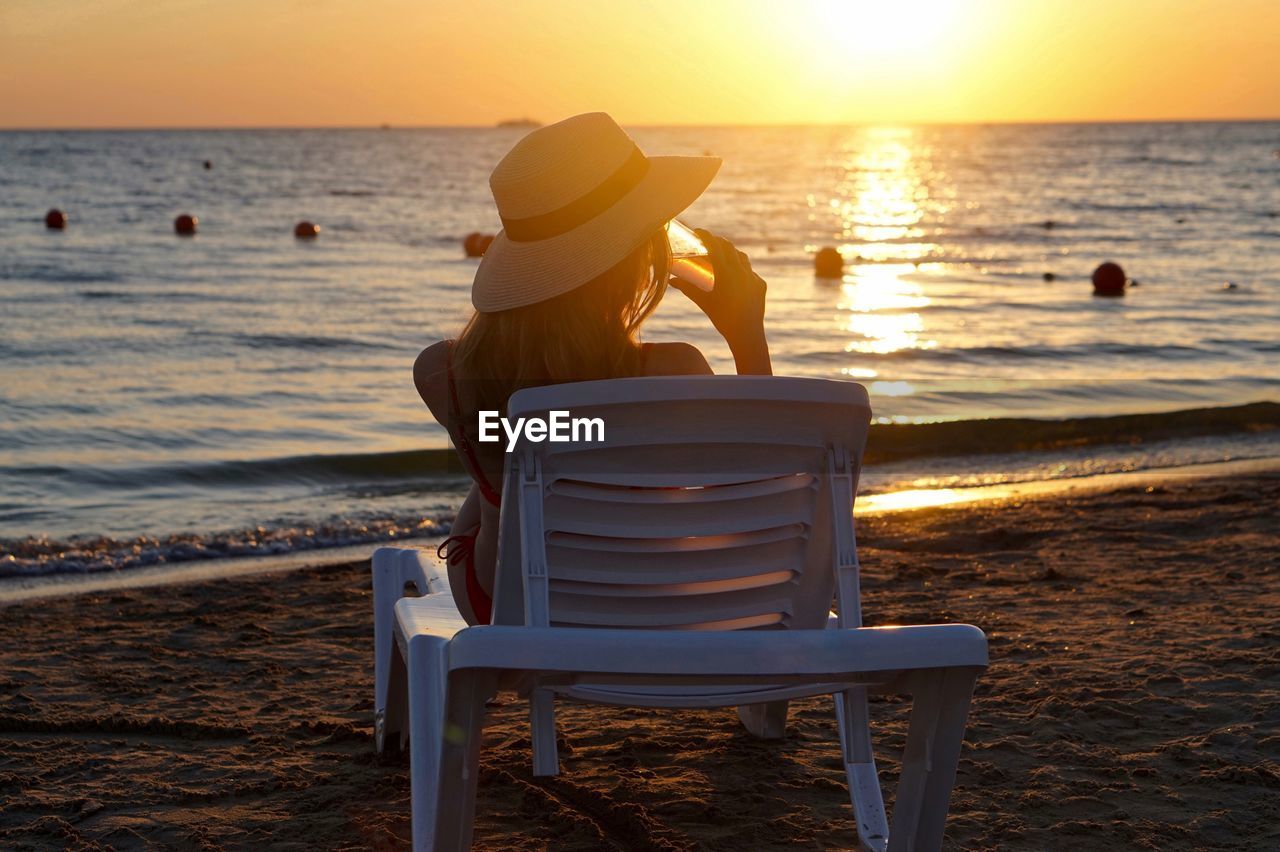 Rear view of woman having drink on lounge chair at beach during sunset