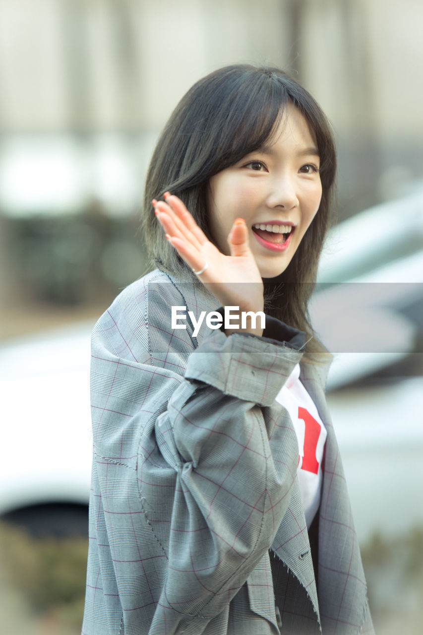 PORTRAIT OF A SMILING YOUNG WOMAN STANDING AGAINST BLURRED BACKGROUND