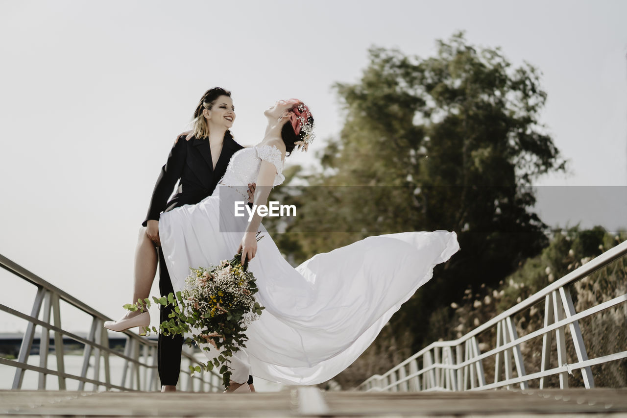 Ground level of cheerful woman in black suit smiling and supporting fiancee in white dress tilting back while standing on bridge on wedding day in park