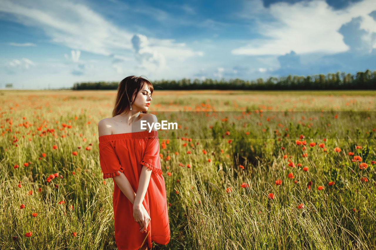 A girl in a red dress above the knee stands in a poppy field during the day against the sky
