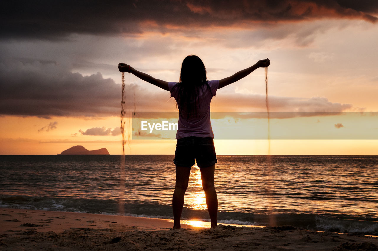 Rear view of silhouette young woman standing at beach against sky during sunset