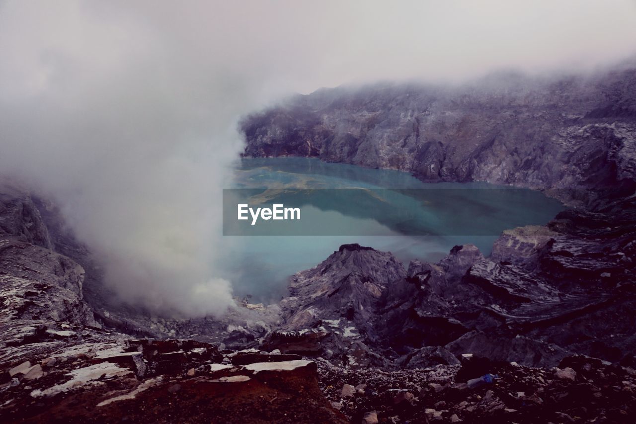 Smoke emitting from volcanic mountain by lake against sky