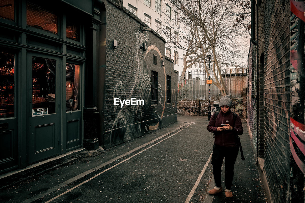 FULL LENGTH OF WOMAN STANDING ON STREET AMIDST BUILDINGS