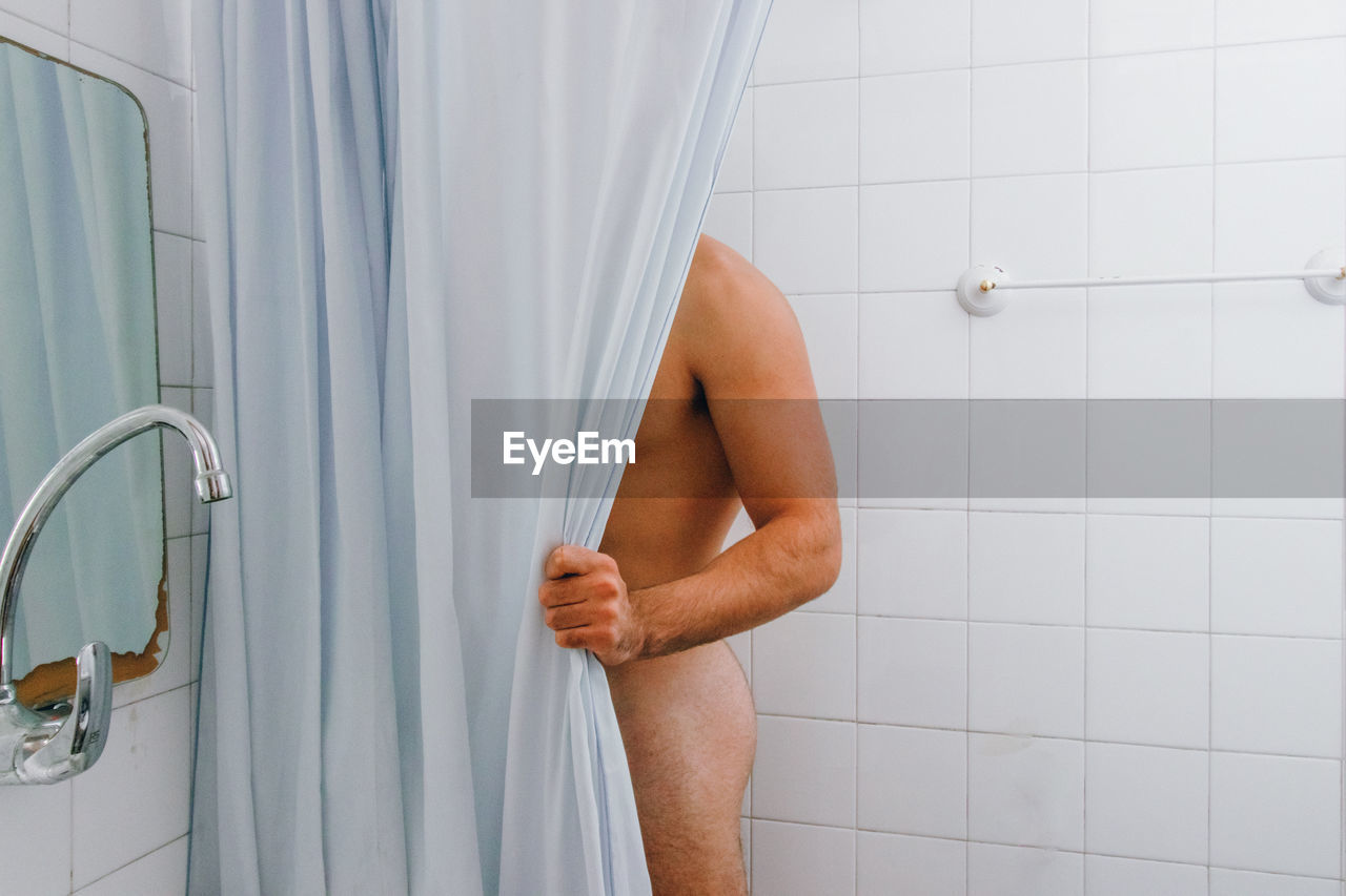 Midsection of naked man standing behind curtain in bathroom at home