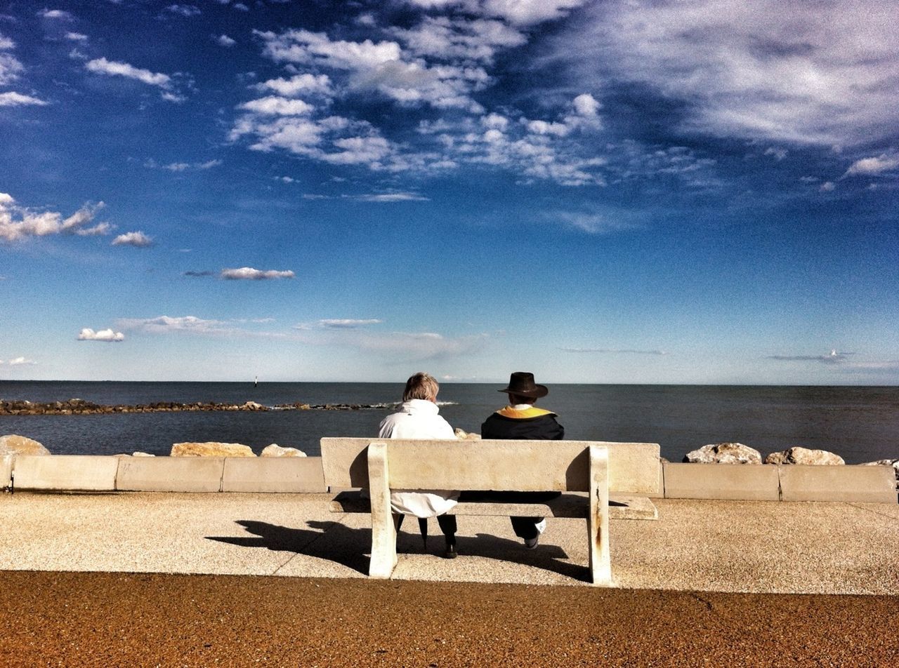 Rear view of people sitting on bench in front of lake against blue sky