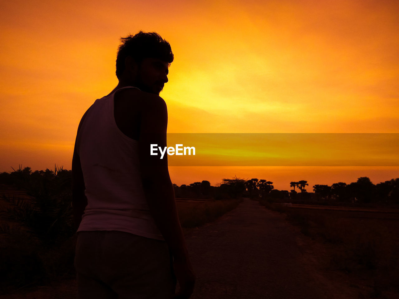 REAR VIEW OF MAN STANDING ON FIELD DURING SUNSET