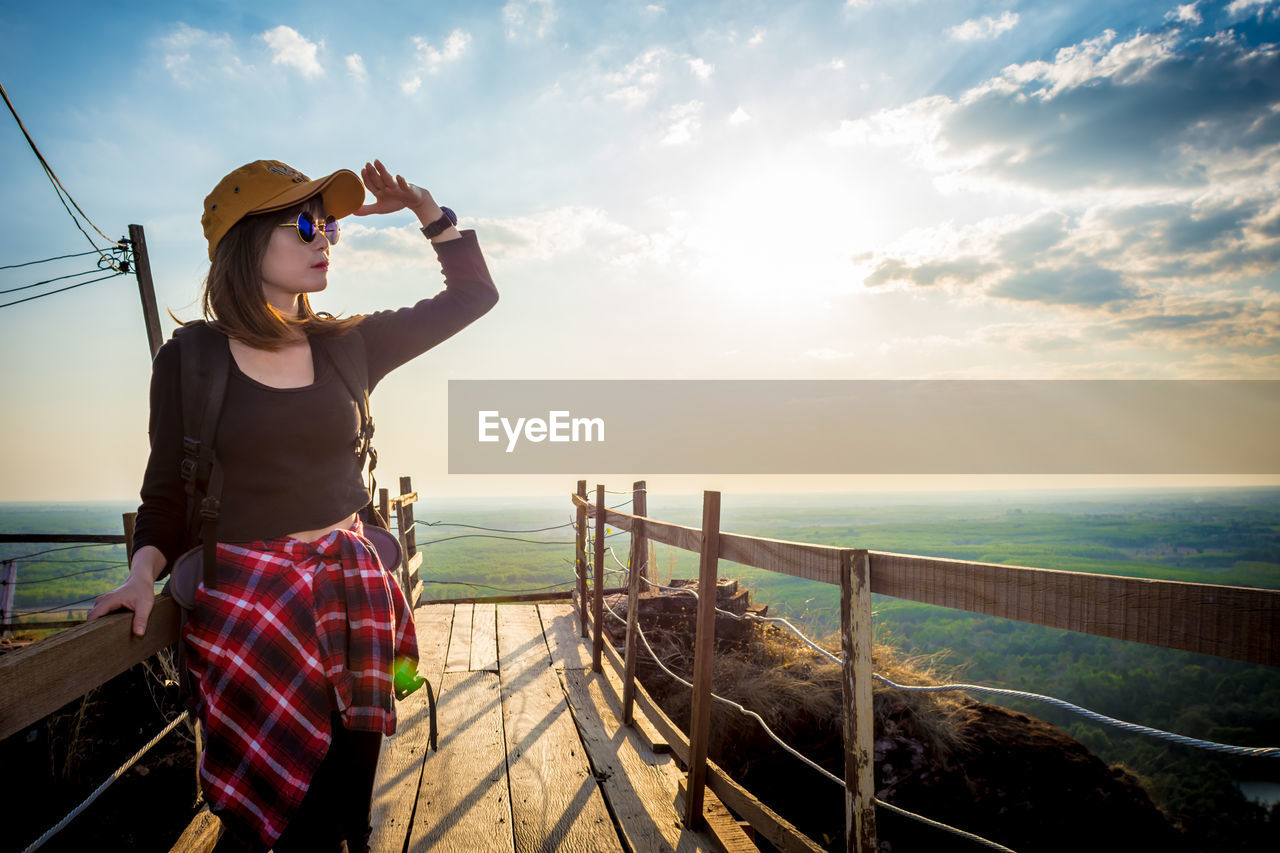 YOUNG WOMAN STANDING BY SEA AGAINST SKY DURING SUNSET