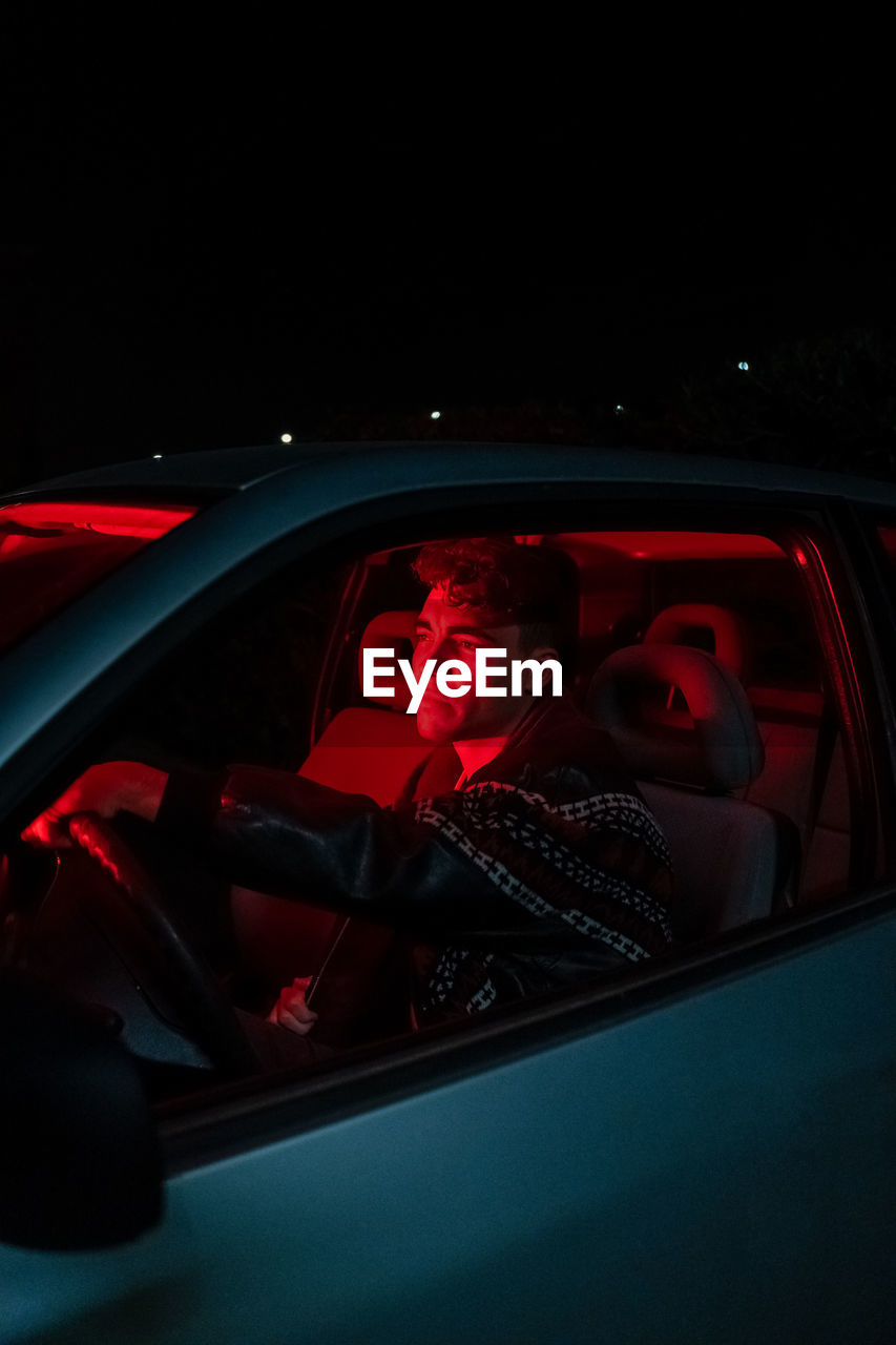 Side view of confident young male millennial sitting in modern car with red neon illumination at night and looking away