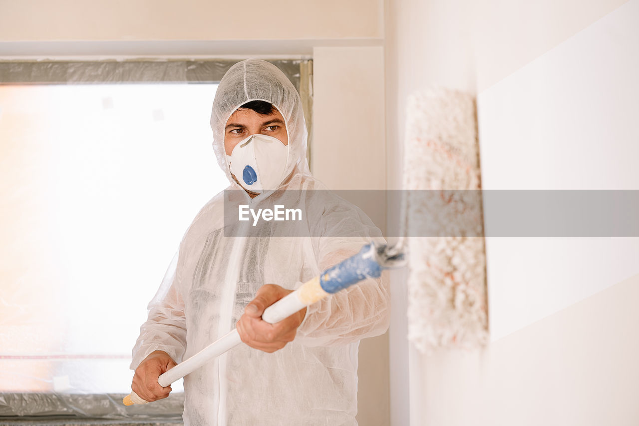 Full length portrait of man working with paint roller in protective suit and mask against wall