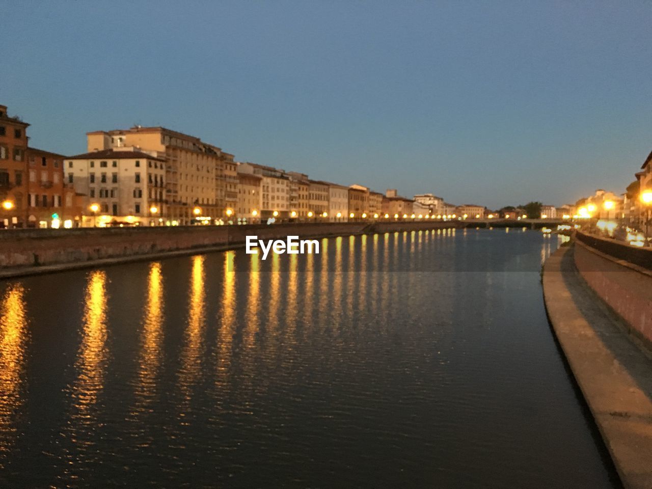 ILLUMINATED BUILDINGS BY RIVER AGAINST CLEAR SKY AT NIGHT
