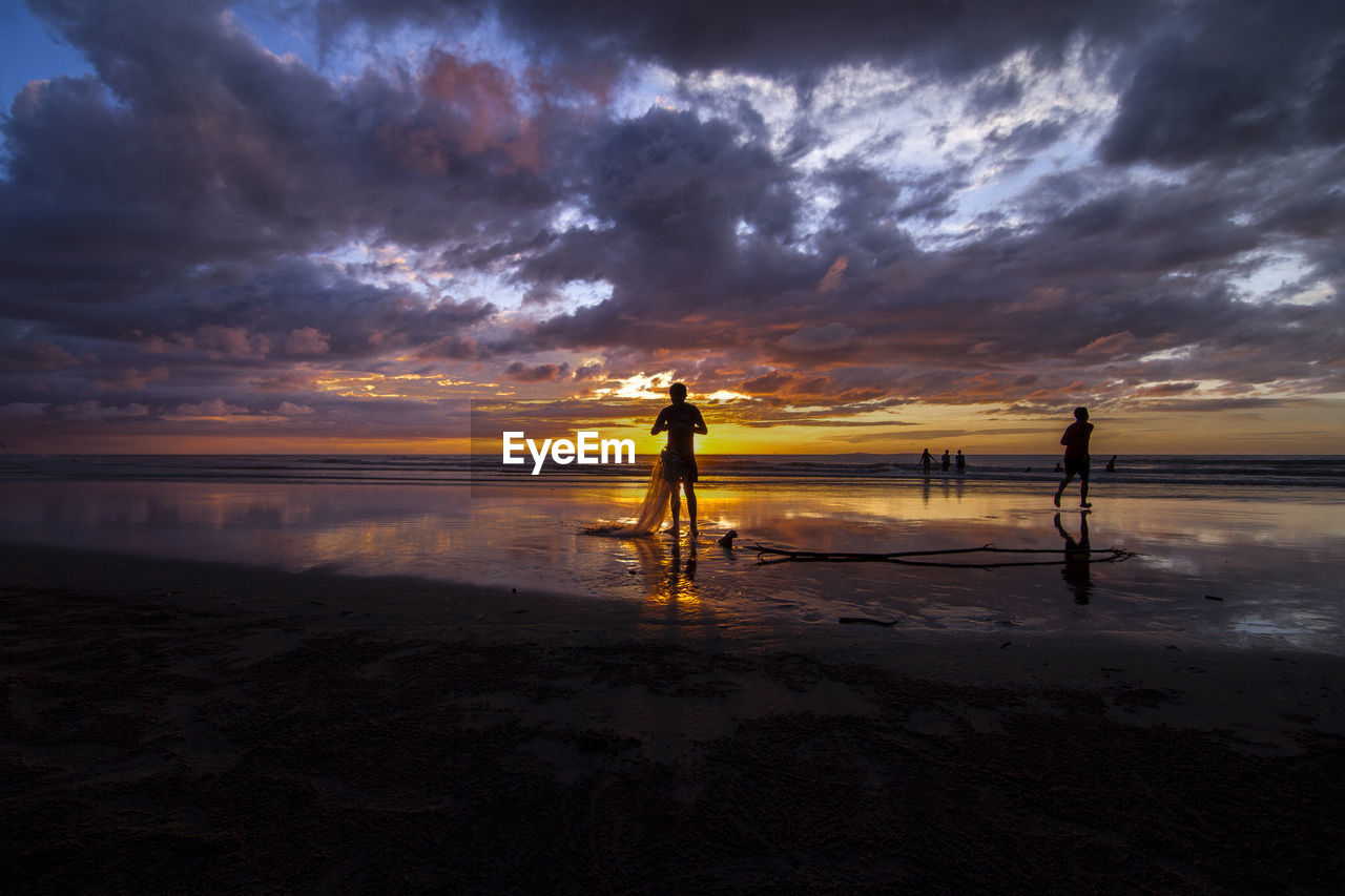 Men standing on beach against cloudy sky during sunset