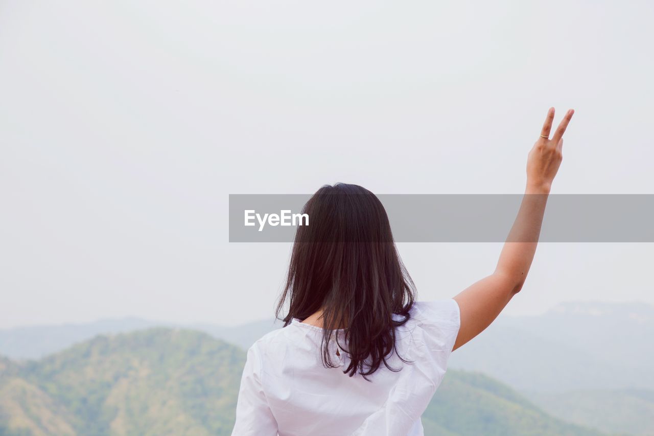 REAR VIEW OF WOMAN WITH ARMS RAISED STANDING ON MOUNTAIN AGAINST SKY