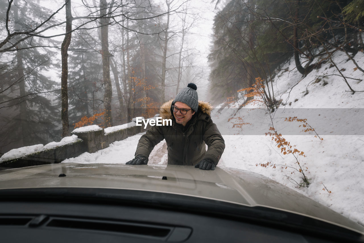 Man pushing car on snow covered road in foggy forest