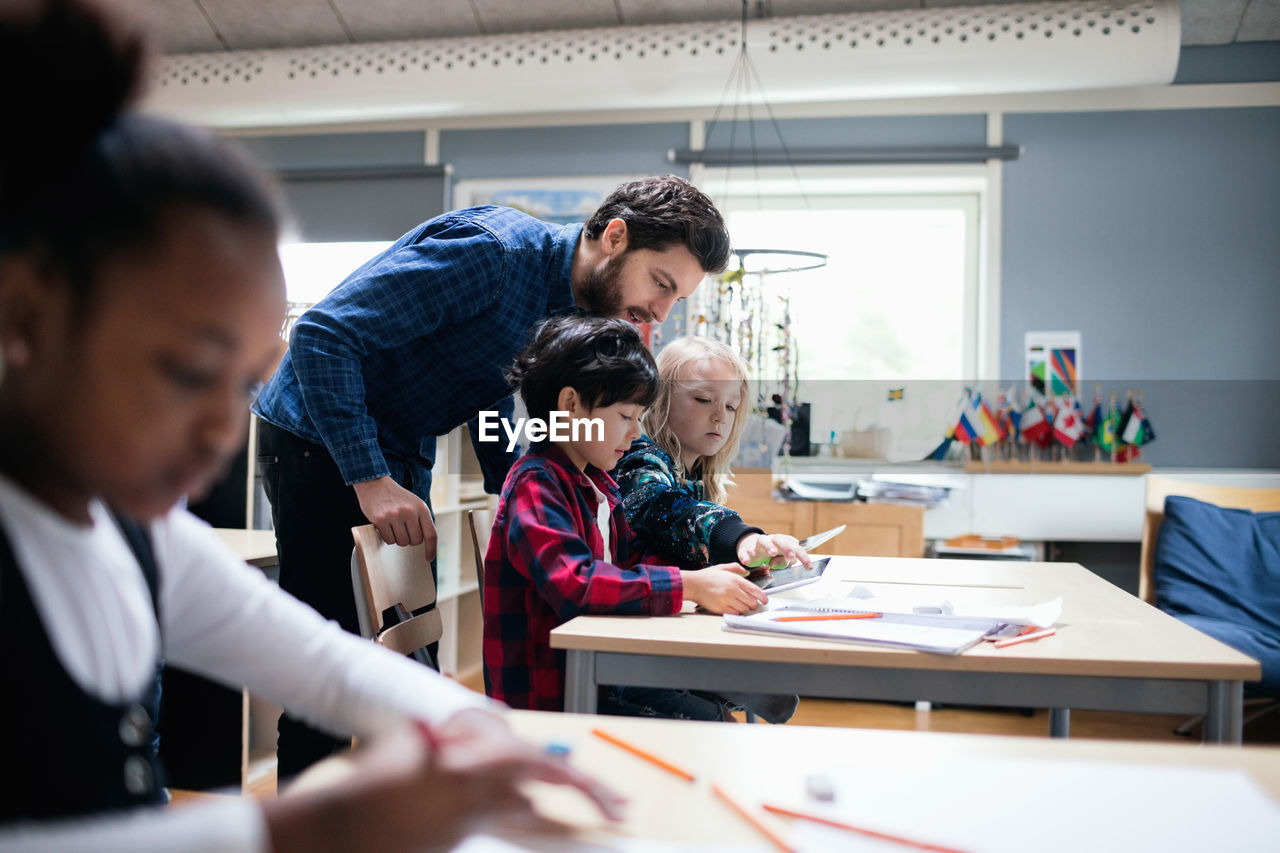 Teacher looking at boy and girl using digital tablet in classroom