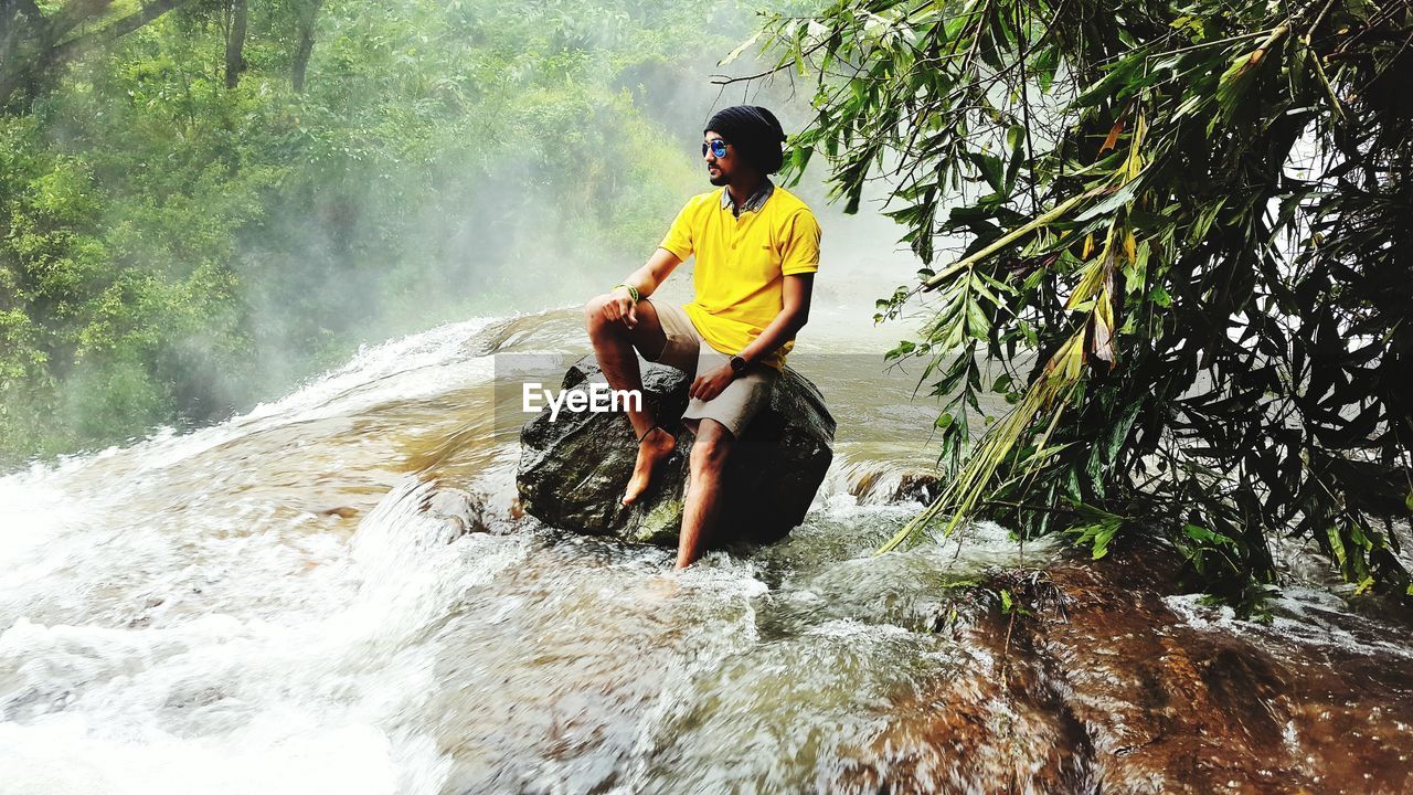 Full length of man sitting on rock in river at forest