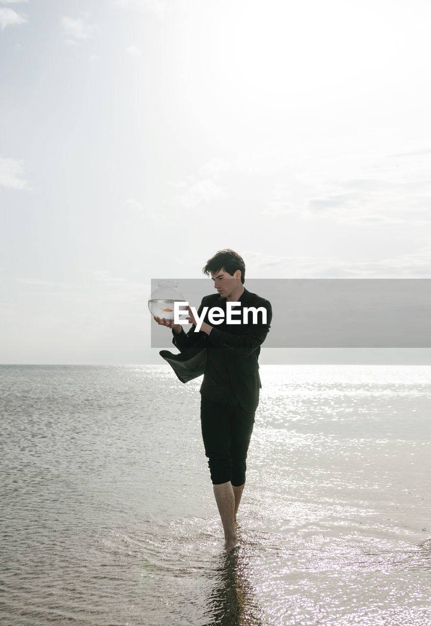 Full length of man holding a goldfish jar  in sea against sky