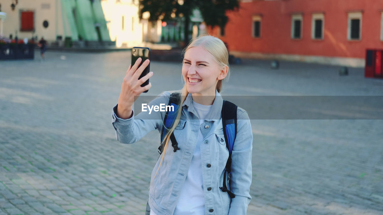 SMILING YOUNG MAN USING PHONE WHILE STANDING ON CITY STREET