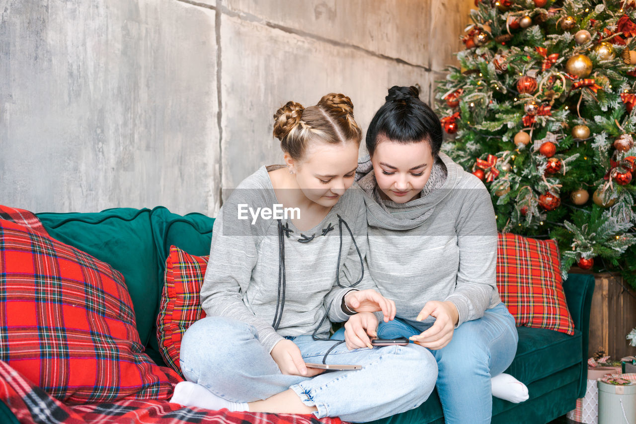 Two pretty young sisters in gray sweatshirts and blue jeans sit on couch near