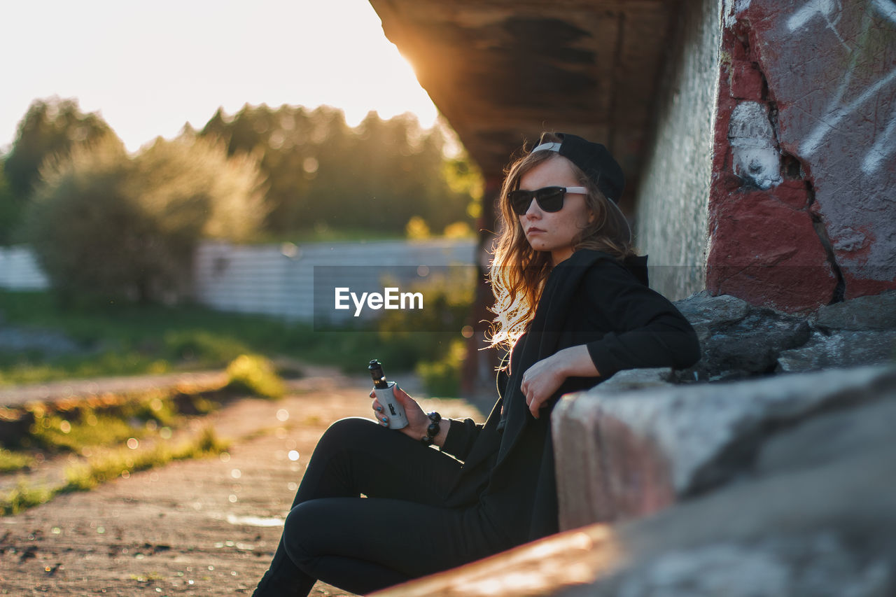 Young woman sitting in sunglasses against sky during sunset