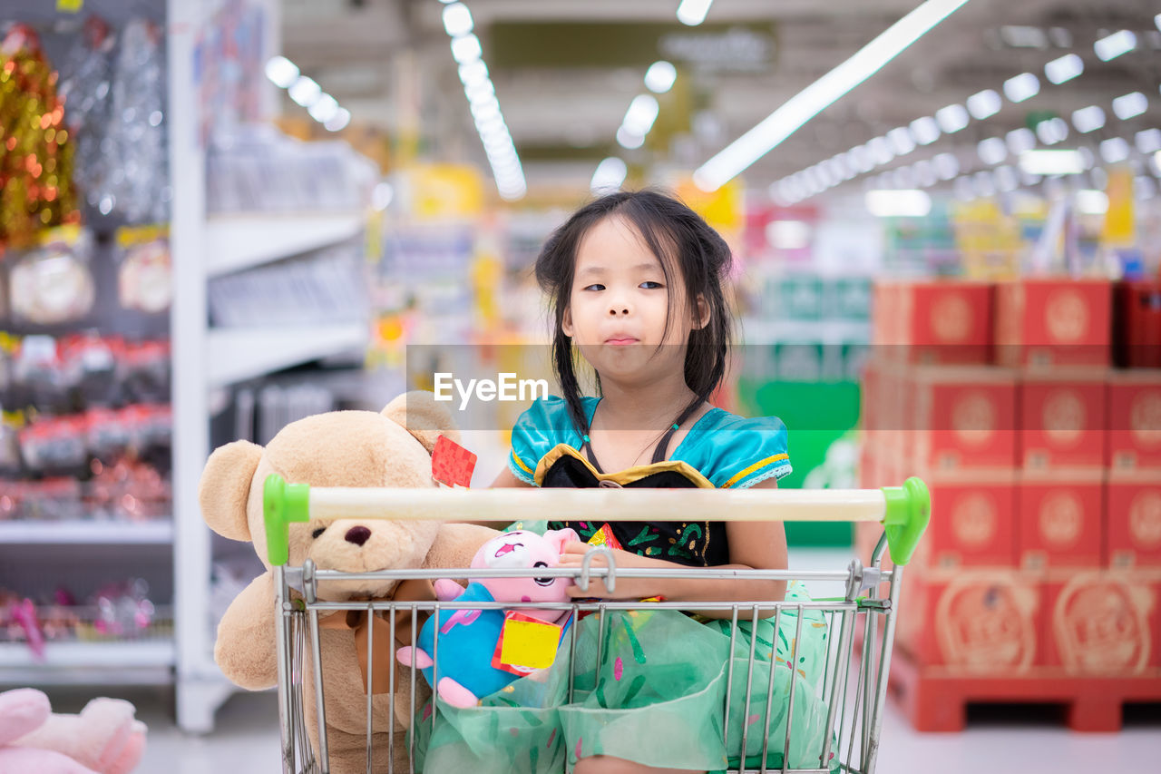 Cute girl looking away while sitting with toys in shopping cart