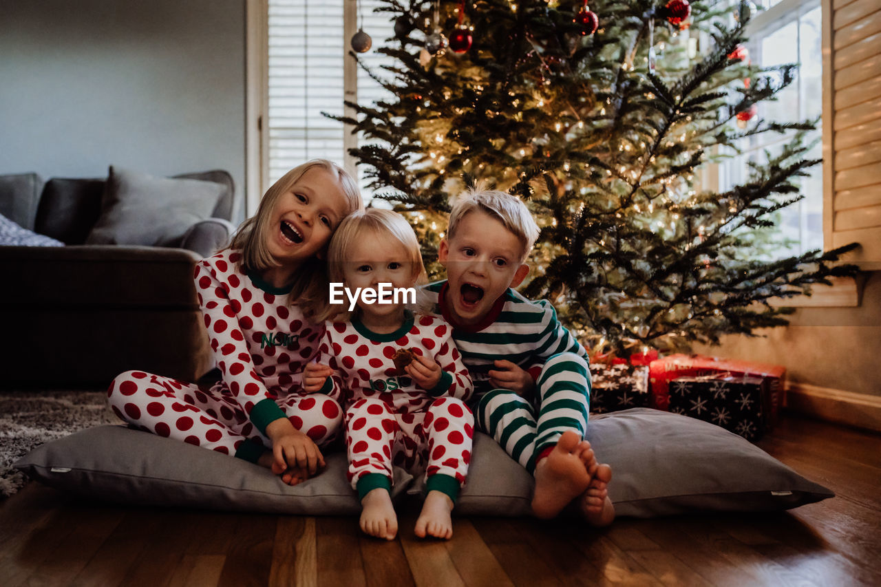 Siblings in costume sitting against christmas tree at home