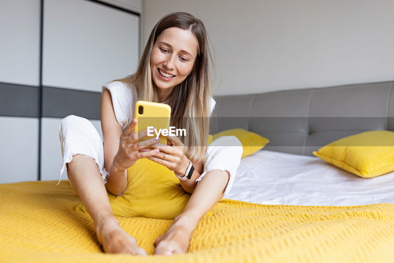 portrait of young woman using mobile phone while sitting on bed at home
