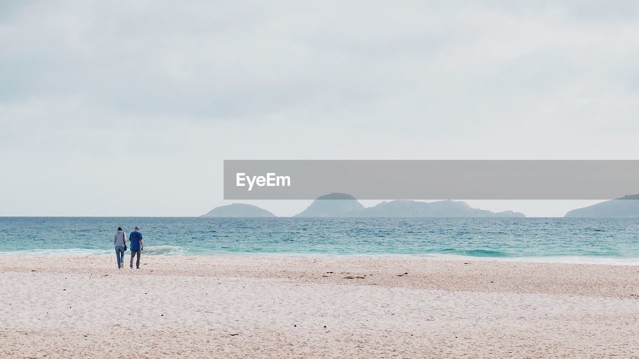 Rear view of couple walking at beach against sky