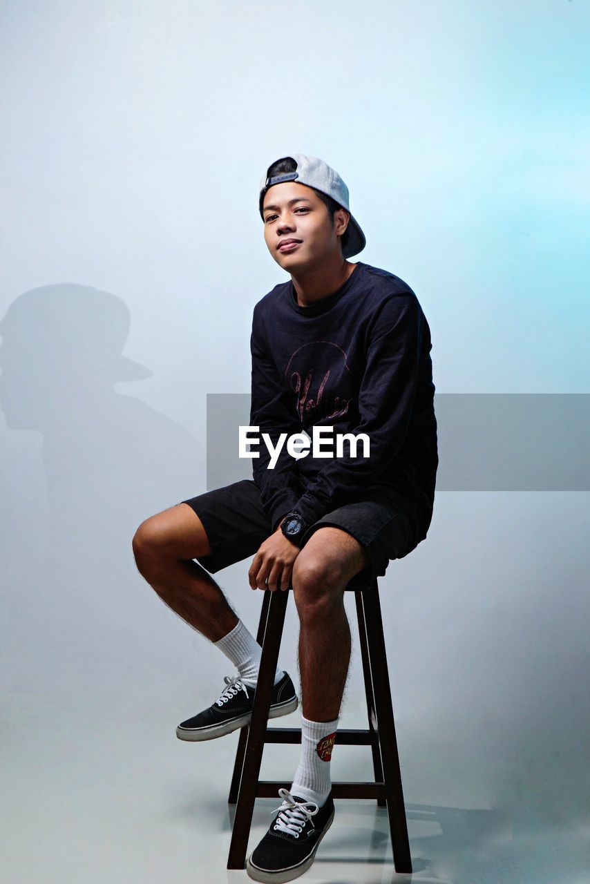 Young man looking away while sitting on seat against wall