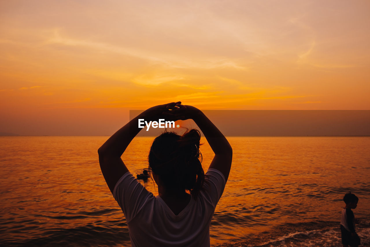 Rear view of woman with arms raised standing by sea against sky at sunset