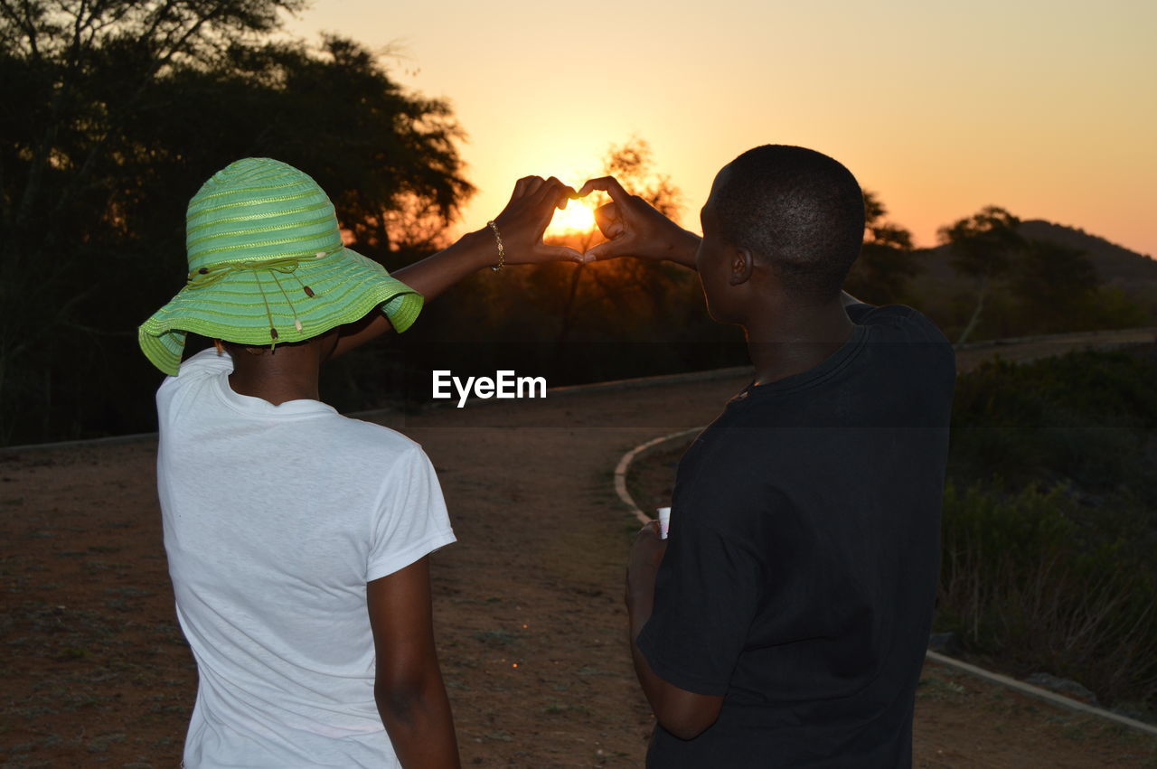 Young man and woman making heart shape against sky during sunset