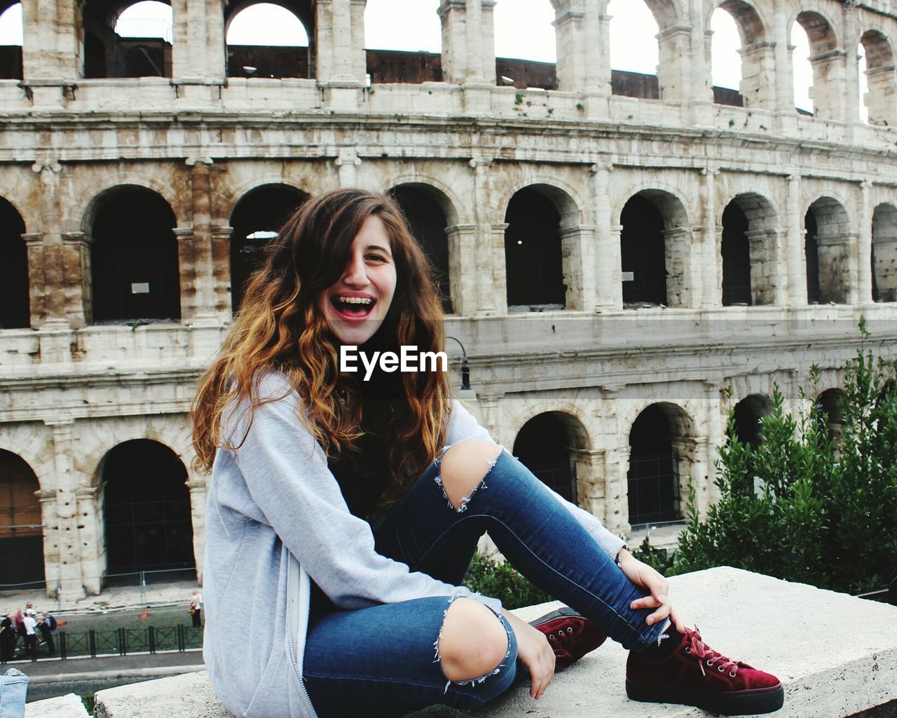 Portrait of happy woman sitting on bench against coliseum