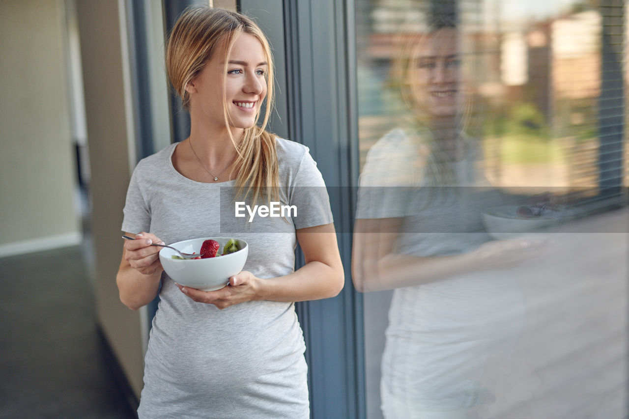 Smiling pregnant young woman having fruit salad by window at home