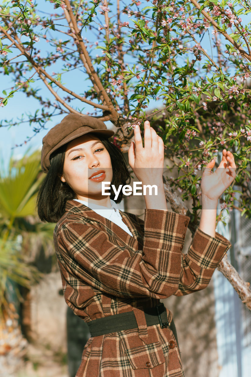 Attractive content brunette wearing trendy checkered coat and hat standing with hands raised in sunny spring garden and looking at camera