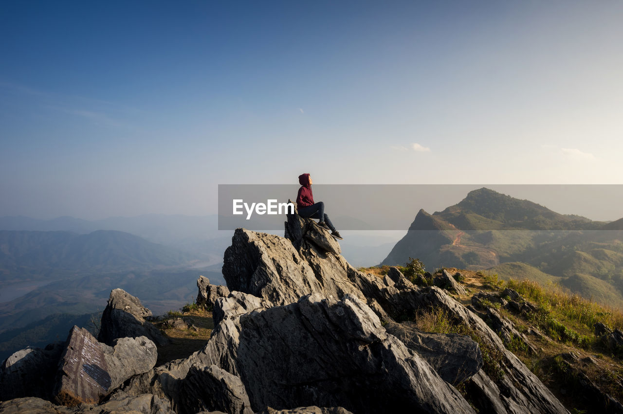 Side view of woman sitting on mountain against blue sky