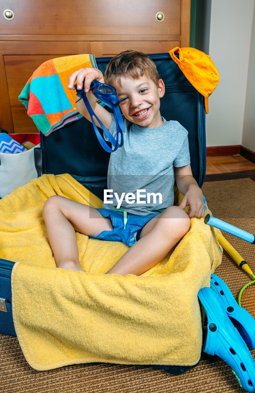 Portrait of smiling boy holding swimming goggles while sitting in suitcase at home