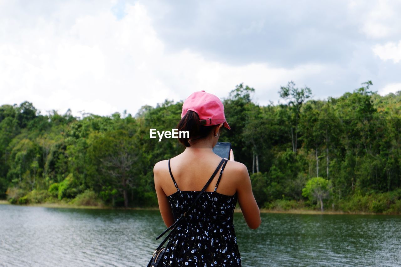WOMAN LOOKING AWAY WHILE STANDING BY LAKE AGAINST TREES