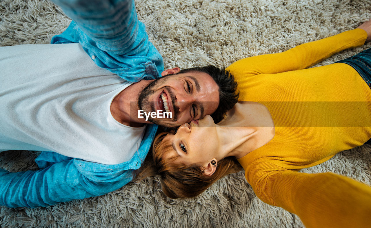 High angle portrait of woman kissing cheerful boyfriend while lying on carpet