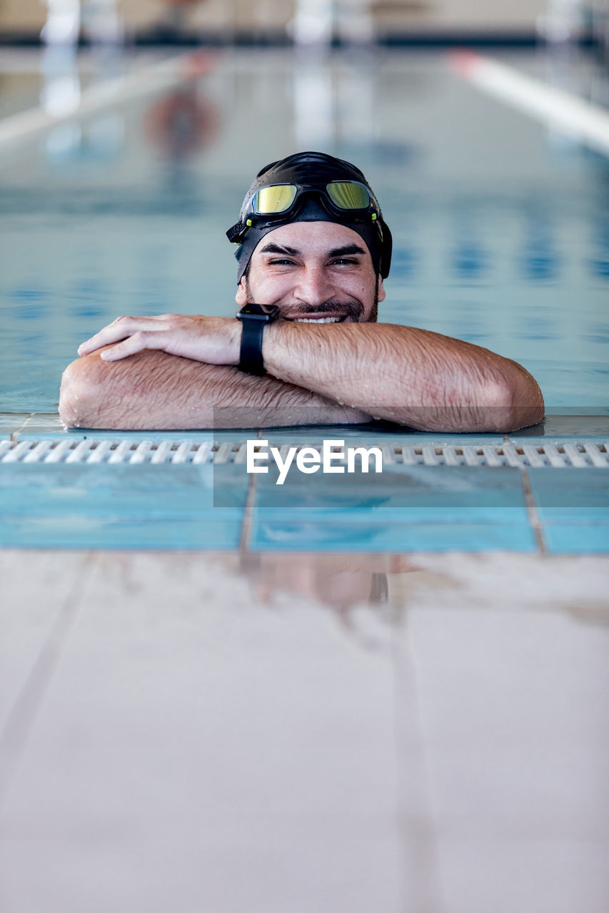 Cheerful sportsman in swimming cap and goggles looking at camera from pool on blurred background