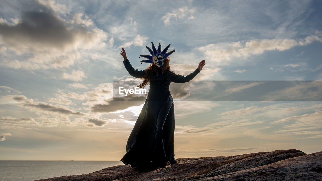Woman wearing mask standing at beach against sky during sunset