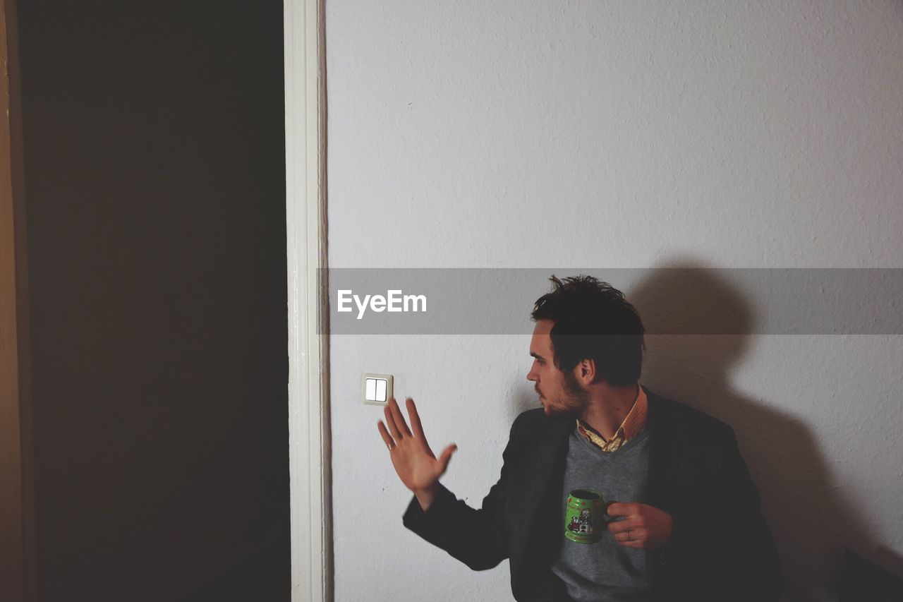 Man gesturing while holding tea cup against wall at home