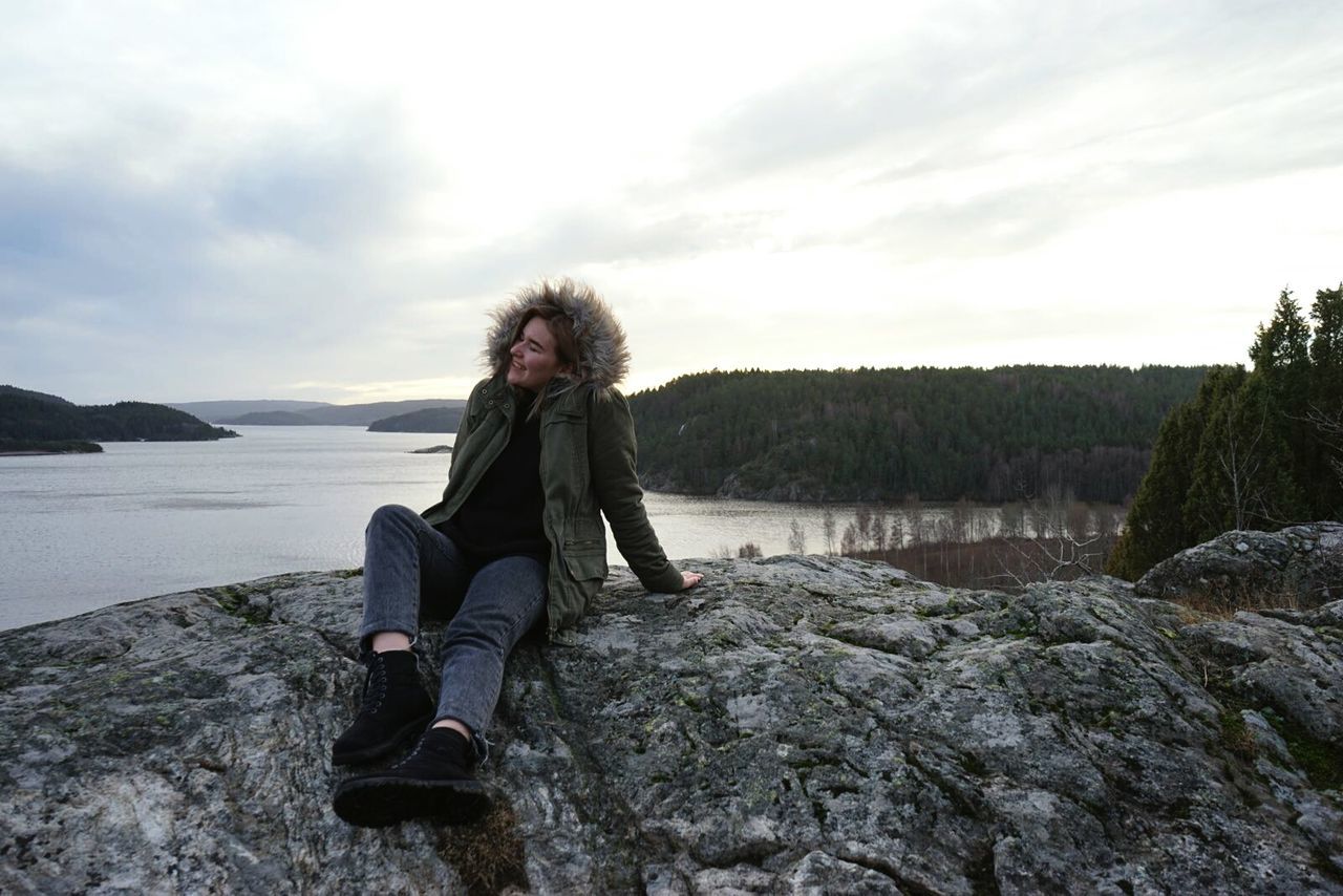 Full length of smiling woman sitting on rock by lake