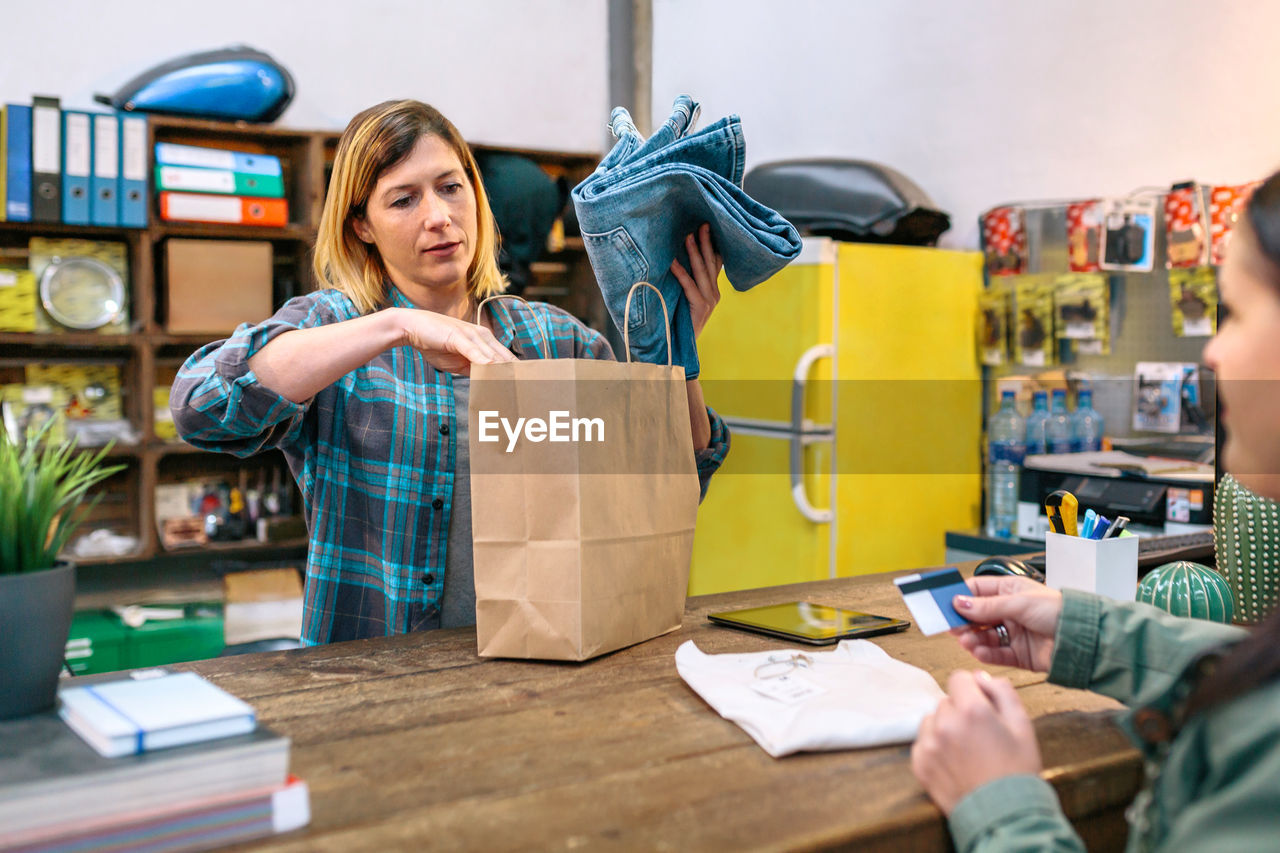 Woman cashier putting jeans into paper bag while female customer holding credit card to pay in store